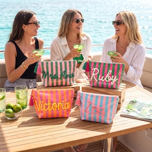 May include: Four rectangular zippered pouches with vertical stripes in various color combinations. Each pouch features a personalized name or title in raised, contrasting script lettering and a matching tassel attached to the zipper pull.