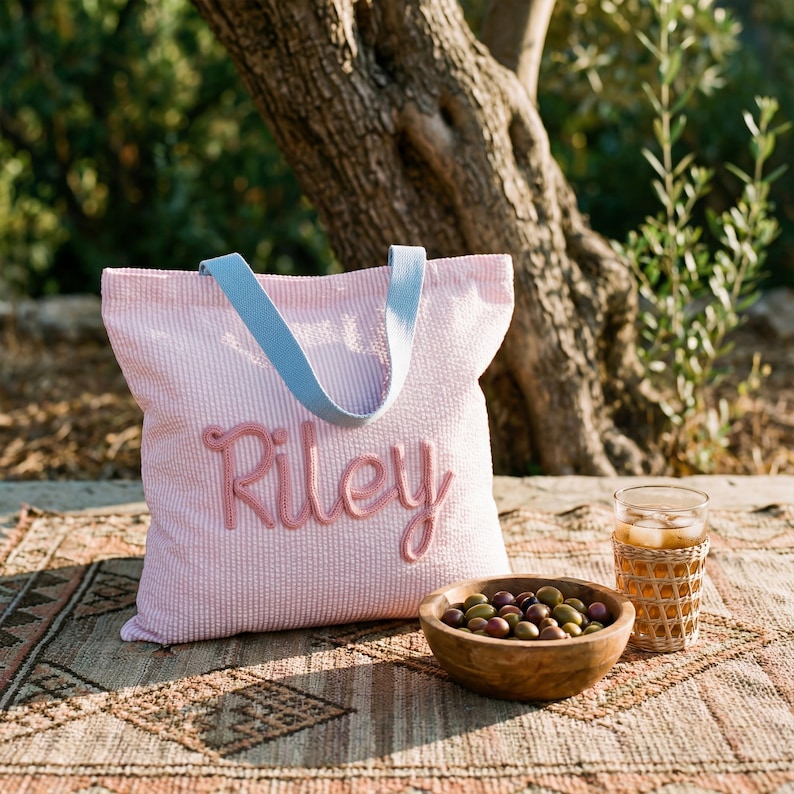 May include: A pink and white striped tote bag with the name "Riley" embroidered in pink script. The bag has light blue handles. A wooden bowl of olives and a glass of iced tea sit nearby.