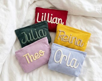 May include: Five rectangular zippered pouches made of ribbed fabric in red, dark green, yellow, light purple, and light blue. Each pouch features a name embroidered in a raised, cursive font: Lillian, Julia, Reina, Theo, and Orla.