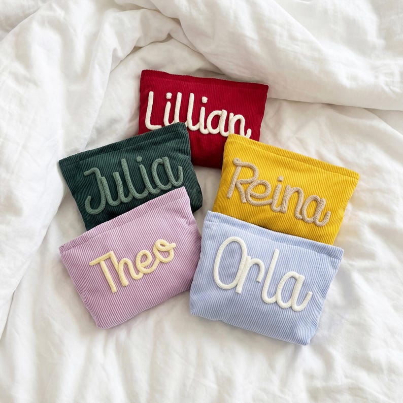 May include: A collection of corduroy pouches in various colors, each embroidered with a name. The pouches are red, green, yellow, lavender, and light blue. Names include "Lillian," "Julia," "Reina," "Theo," and "Orla" in a cursive font.