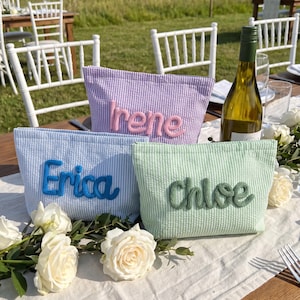 May include: Three seersucker fabric pouches in light blue, lavender, and mint green. Each pouch features a name embroidered in raised yarn: Erica in blue, Irene in pink, and Chloe in dark green. The pouches are arranged on a white table runner.