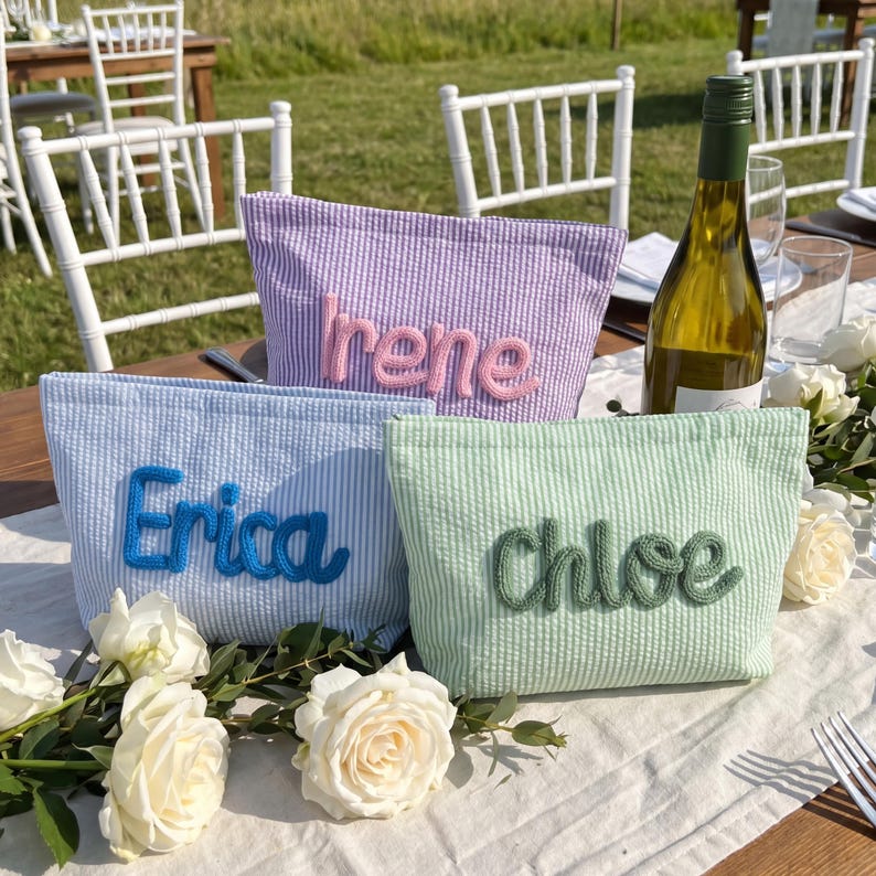 May include: Three seersucker fabric pouches in light blue, lavender, and mint green. Each pouch features a name embroidered in raised yarn: Erica in blue, Irene in pink, and Chloe in dark green. The pouches are arranged on a white table runner.