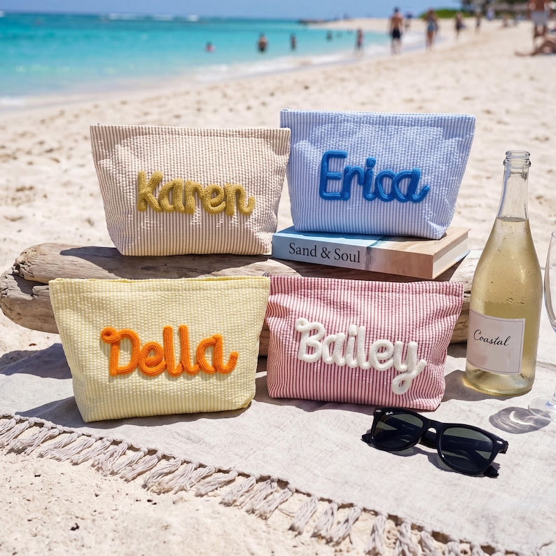 May include: Four seersucker pouches in tan, light blue, yellow, and red stripes. Each pouch features a name embroidered in raised script: Karen in yellow, Erica in blue, Della in orange, and Bailey in white. A bottle of Coastal wine and black sunglasses.
