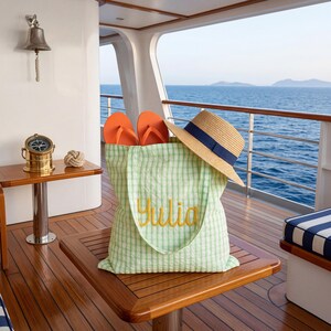 Pode incluir: Uma sacola verde e branca com o nome "Julia" bordado em amarelo, contendo chinelos laranja e um chap&eacute;u de palha com uma faixa azul. Os itens est&atilde;o em uma mesa de madeira em um barco, com o mar ao fundo.
