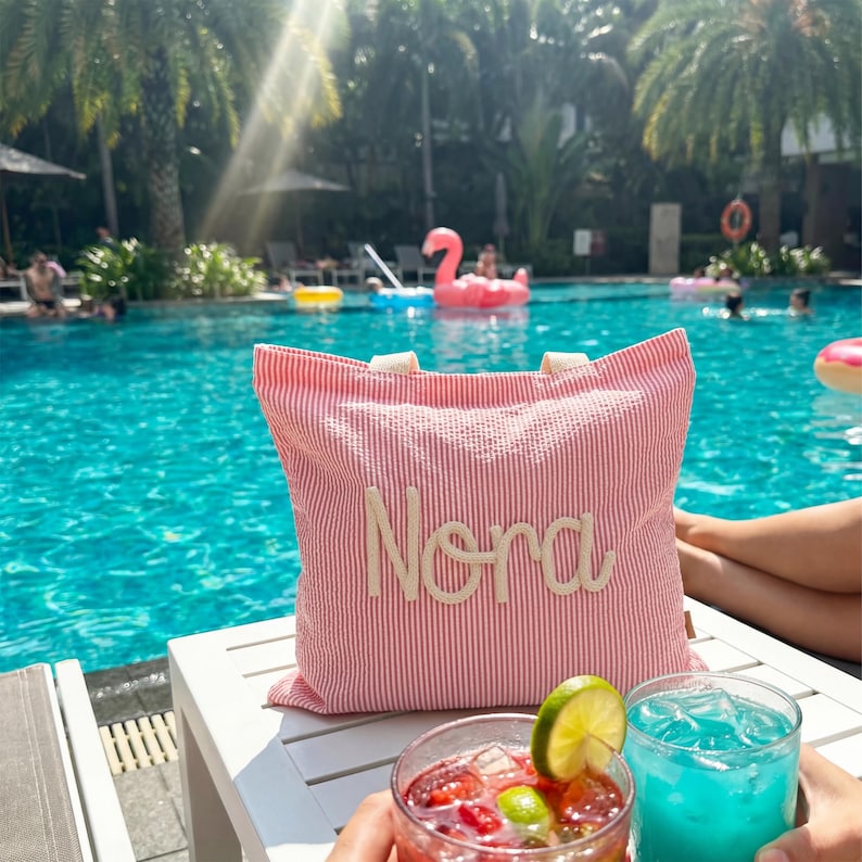 May include: A red and white striped tote bag with the name "Nora" embroidered in cream-colored thread. The bag has cream-colored handles and is sitting on a white table next to two cocktails. The background is a swimming pool.