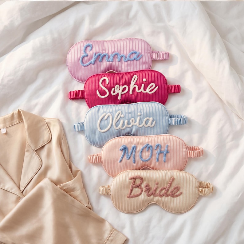 May include: Five oval-shaped sleep masks with a vertical striped texture and elastic headbands. The masks feature embroidered names and titles: Emma in blue, Sophie in white, Olivia in white, MOH in blue, and Bride in dark pink thread.