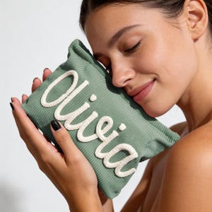 May include: A sage green corduroy pillow with the name "Olivia" embroidered in white script. The pillow is rectangular and appears soft. The image is a close-up, highlighting the texture and design of the personalized item.