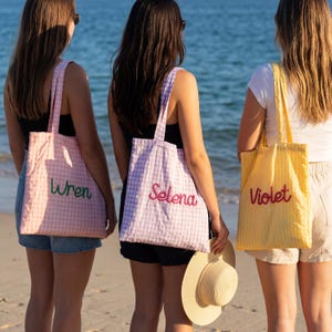 Pode incluir: Tr&ecirc;s sacolas com nomes bordados: "Wren" em verde em uma sacola rosa xadrez, "Selena" em vermelho em uma sacola roxa xadrez e "Violet" em vermelho em uma sacola listrada amarela. As sacolas t&ecirc;m al&ccedil;as combinando.