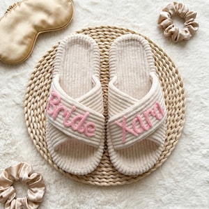 Könnte beinhalten: Ein Paar cremefarbene Cord-Hausschuhe mit überkreuzten Riemen, die den rosa gestickten Text Bride auf dem linken und Xara auf dem rechten Schuh tragen, dazu eine passende cremefarbene Schlafmaske aus Satin und zwei passende Satin-Haargummis.