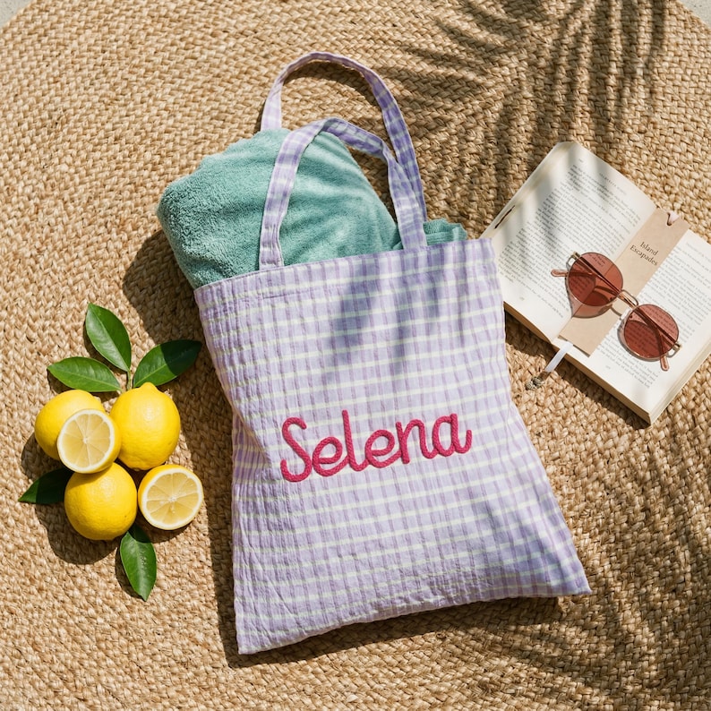 Pode incluir: Uma sacola tote xadrez lil&aacute;s e branca com o nome "Selena" bordado em rosa. A sacola cont&eacute;m uma toalha verde dobrada. Lim&otilde;es, um livro e &oacute;culos de sol est&atilde;o ao lado da sacola.