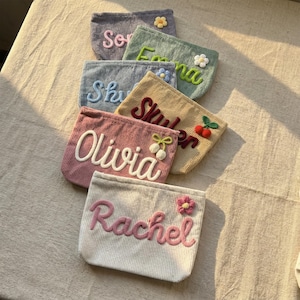 May include: A collection of corduroy pouches in various pastel colors, each embroidered with a name in a contrasting color. Some pouches feature floral or fruit embellishments. The pouches are arranged on a textured surface.