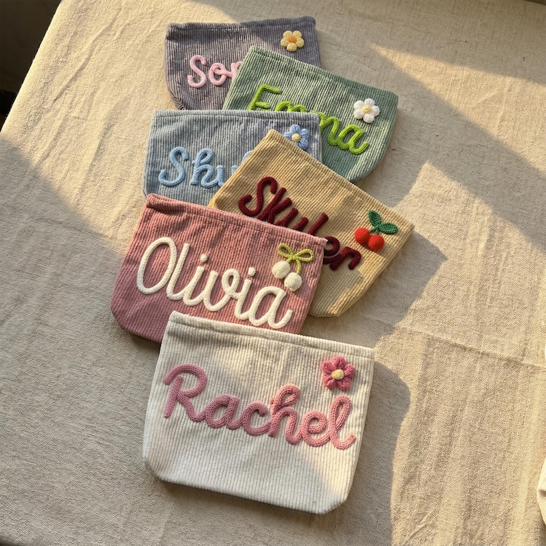 May include: A collection of corduroy pouches in various pastel colors, each embroidered with a name in a contrasting color. Some pouches feature floral or fruit embellishments. The pouches are arranged on a textured surface.