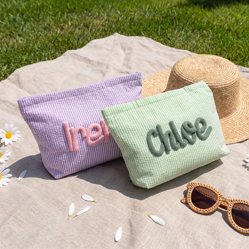 May include: Two seersucker cosmetic bags, one lavender with "Ire" in pink, the other mint green with "Chloe" in green. A straw hat and sunglasses rest on a neutral-toned blanket on grass.
