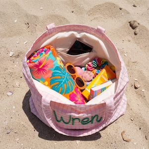 Pode incluir: Uma sacola rosa xadrez cheia de itens essenciais para a praia, incluindo uma toalha floral enrolada, &oacute;culos de sol laranja, protetor solar e um livro. A sacola tem o nome "Wren" bordado em verde na frente. A sacola est&aacute; em uma praia arenosa.