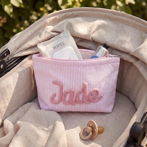 May include: A light pink and white seersucker zippered pouch featuring the name Jade in raised pink yarn lettering. The pouch contains a pack of wipes and a metal tube, with a beige silicone pacifier resting in front of the fabric accessory.