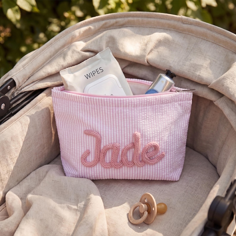 May include: A light pink and white seersucker zippered pouch featuring the name Jade in raised pink yarn lettering. The pouch contains a pack of wipes and a metal tube, with a beige silicone pacifier resting in front of the fabric accessory.