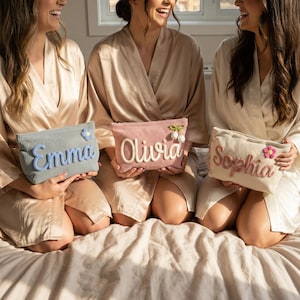 May include: Three women holding personalized corduroy cosmetic bags. The bags are in light blue, pink, and cream colors, each embroidered with a name in a complementary color. The bags are decorated with floral or pom-pom accents.