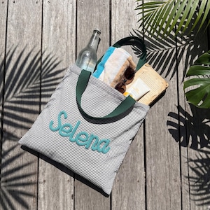May include: A gray and white striped tote bag with the name "Selena" embroidered in teal. The bag has green handles and is filled with a towel, sunglasses, sunscreen, a book, and a glass bottle.