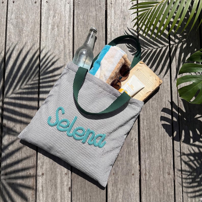 May include: A gray and white striped tote bag with the name "Selena" embroidered in teal. The bag has green handles and is filled with a towel, sunglasses, sunscreen, a book, and a glass bottle.