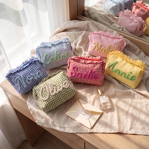 May include: A collection of rectangular fabric pouches featuring a gingham pattern in various colors including blue, green, pink, red, and yellow. Each pouch has a ruffled top edge and a personalized name embroidered in a textured script font.