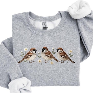 May include: A grey sweatshirt with a crew neck and white fleece lining. The front displays an embroidered design of three brown sparrows amongst white daisies. The sleeves are cuffed. The garment is a comfortable, casual style.