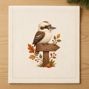 May include: A square off-white fabric panel featuring an embroidered kookaburra perched on a wooden signpost. The bird has brown and cream feathers with a blue wing patch. The sign is surrounded by embroidered autumn leaves and small red berries.
