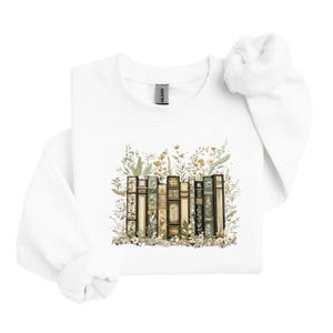 May include: White sweatshirt featuring a watercolor illustration of a row of vintage books. The books are in various colors, including green, gold, and brown, with floral accents. The design is centered on the front of the sweatshirt.