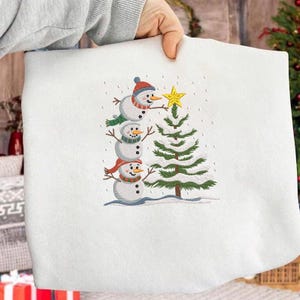 Op de afbeelding: Wit sweatshirt met een geborduurd ontwerp van drie op elkaar gestapelde sneeuwmannen, met hoeden en sjaals. Een kerstboom met een gele ster staat naast de sneeuwmannen. Sneeuwvlokken vallen.