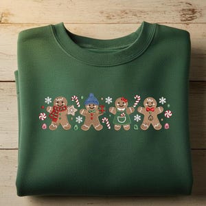 Könnte beinhalten: Ein gefaltetes, dunkelgrünes Sweatshirt mit einem gestickten Design von vier Lebkuchenmännchen. Jedes Lebkuchenmännchen hat einzigartige Details, darunter Hüte, Schals und Zuckerstangen. Schneeflocken und andere festliche Elemente vervollständigen das Weihnachtsthema.