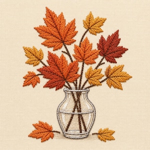 May include: An embroidery design on cream fabric featuring a clear glass vase filled with autumn maple leaves in shades of orange, red, and yellow. Two additional fallen leaves are positioned at the base of the vase on the fabric surface.