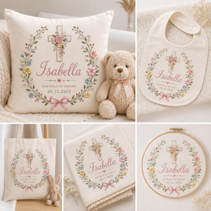 May include: A collection of personalized baptism gifts, including a pillow, bib, tote bag, handkerchief, and embroidery hoop. Each item features the name "Isabella", a cross, floral wreath, and the date "05.12.2025". A teddy bear and bunny are also present.
