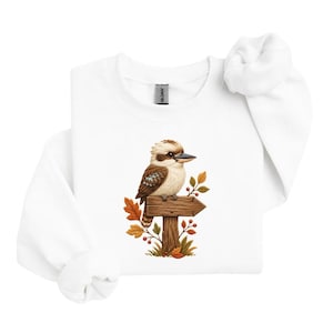 May include: A white crewneck sweatshirt featuring a central graphic of a brown and white kookaburra perched on a wooden signpost. The sign is surrounded by autumn-colored leaves in shades of orange, yellow, and brown, along with small red berries.