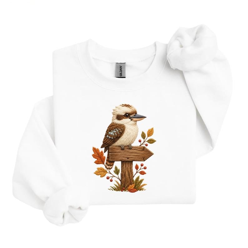 May include: A white crewneck sweatshirt featuring a central graphic of a brown and white kookaburra perched on a wooden signpost. The sign is surrounded by autumn-colored leaves in shades of orange, yellow, and brown, along with small red berries.