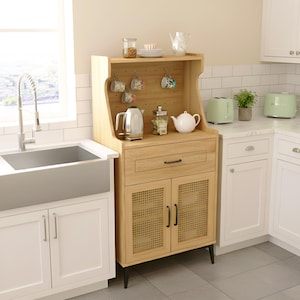 May include: A light wood kitchen hutch with a cabinet, drawer, and shelf. It features a woven cane design on the cabinet doors and black legs. The shelf holds a teapot, tea cups, and a kettle. The kitchen has white cabinets and a stainless steel sink.