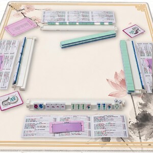 May include: A complete Mahjong set with a cream-colored playing surface featuring floral and botanical designs. The set includes tiles, racks, and score cards. The tiles are primarily green and white, with some tiles displaying colorful symbols.