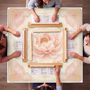 May include: A square Mahjong table with a floral design and four players. The table features a large, light pink peony in the center, surrounded by a cream-colored border with gold accents. Wooden racks hold tiles, and players are seated around the table.