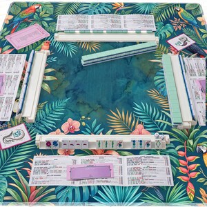 May include: A Mahjong set with white and green tiles, dice, and scorecards, arranged on a tropical-themed mat with parrots and floral patterns. The set includes racks and a variety of tiles for gameplay.