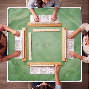 May include: A green Mahjong game table with a marble-like design, surrounded by four players. The table has wooden racks and tiles, with the word "NAME" in elegant script. The players are arranging their tiles and cards.