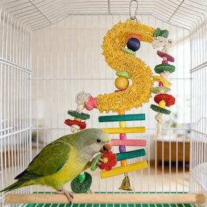 Puede incluir: Un colorido juguete para pájaros dentro de una jaula. El juguete presenta una estructura amarilla en forma de S con varios elementos de madera y cuerda, incluyendo una campana. Un pequeño pájaro verde y amarillo está posado en una varilla de madera, interactuando con el juguete.