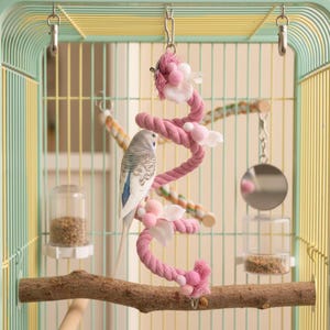 May include: A budgie perches on a wooden branch inside a birdcage. The cage features a pink rope toy with white flower decorations. The cage is painted in shades of yellow and green. A mirror and food containers are also visible.
