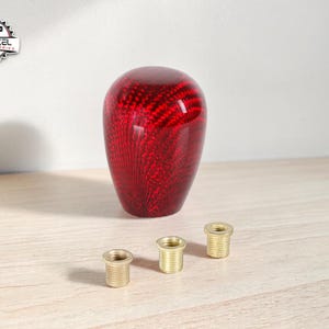 May include: A red carbon fibre shift knob with a glossy finish. Three brass threaded inserts are positioned below the knob. The logo "YEHEZKEL CAR ACCESSORIES" is in the upper left corner.