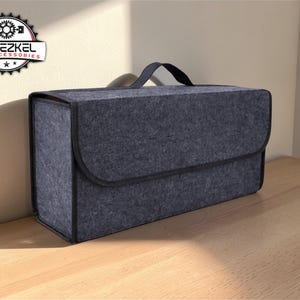 May include: A dark gray felt car organizer with a black handle and a curved flap closure. The organizer has a rectangular shape and is designed for car accessories. The logo "YEHEZKEL CAR ACCESSORIES" is visible in the upper left corner.