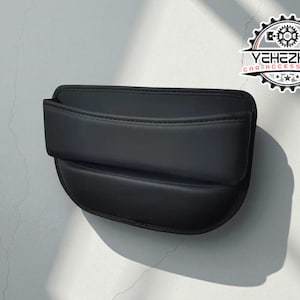 May include: Black car seat organiser with a curved design and a stitched seam. The organiser has a pocket and is made of a smooth material. The logo "YEHEZKEL CAR ACCESSORIES" is visible in the upper right corner.