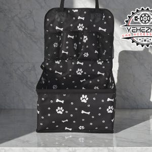 Könnte beinhalten: Schwarzer Auto-Organizer mit weißen Pfotenabdrücken und Knochenmustern. Verfügt über mehrere Taschen und eine Aufbewahrungsbox. Enthält einen Griff zum einfachen Tragen. Das Logo "YEHEZKEL CAR ACCESSORIES" ist sichtbar.