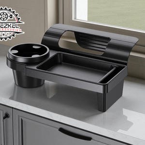May include: A black car accessory organizer with a cup holder, tray, and slot. The organizer is made of plastic and sits on a countertop near a window. The logo "YEHEZKEL CAR ACCESSORIES" is visible.
