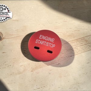 Op de afbeelding: Een rode start/stop-knop voor de motor van een auto met de witte tekst "ENGINE START/STOP". De knop heeft twee kleine rechthoekige sleuven en ligt op een lichte houtnerf. Een logo is zichtbaar in de linkerbovenhoek.