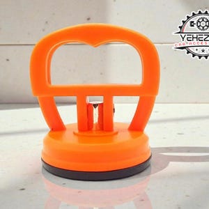 Könnte beinhalten: Ein orangefarbener Saugnapf-Dellenentferner mit schwarzem Sockel und herzförmigem Griff. Das Werkzeug hat eine kreisförmige Basis und dient zum Entfernen von Dellen an Fahrzeugen. Das Logo "YEHEZKEL CAR ACCESSORIES" ist im Hintergrund sichtbar.