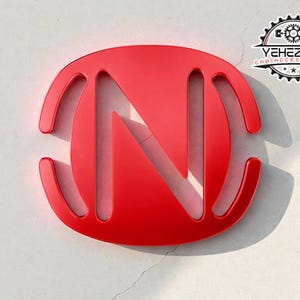 Może przedstawiać: Błyszczące czerwone logo z literą "N" w owalnym kształcie, z zakrzywionymi liniami po bokach. Logo umieszczone jest na białym, teksturowanym tle. Tekst "YEHEZKEL CAR ACCESSORIES" znajduje się w czarno-białym okrągłym logo.