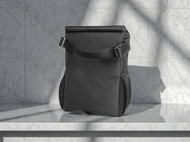 Peut inclure: Un sac &agrave; d&eacute;jeuner isotherme noir avec une bandouli&egrave;re et une poche lat&eacute;rale en filet. Le sac a une fermeture &agrave; enroulement et une boucle. Le sac est con&ccedil;u pour garder les aliments et les boissons au frais. Le sac est pos&eacute; sur une surface blanche.