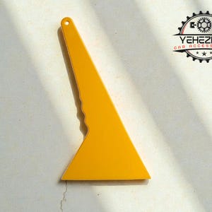 Könnte beinhalten: Ein gelber Auto-Zubehörartikel, ein Fensterschaber, wird auf einer weißen Oberfläche gezeigt. Der Schaber hat eine dreieckige Form mit einem Griff und einer gebogenen Kante. Das Logo "YEHEZKEL CAR ACCESSORIES" ist oben rechts sichtbar.